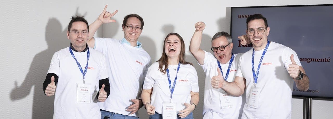 A group of five diverse professionals, four men and one woman, are captured in a moment of high-energy celebration during an AI hackathon. They are all wearing matching white t-shirts with red branding and blue lanyards with ID badges. Their expressions are joyful, with wide smiles and cheers. They are gesturing with thumbs-up and rock-on hand signs against a clean white background, with a digital screen partially visible on the right.