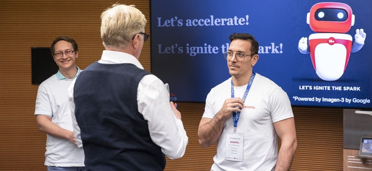 A dynamic scene from an AI hackathon featuring three professional men in conversation. The central figure, seen from behind, wears a navy sweater vest over a crisp white shirt. Facing him is a man in a white t-shirt and glasses, holding a blue lanyard and looking focused. In the background, a third man smiles warmly. A large digital display shows the text "Let's accelerate! Let's ignite the spark!" alongside a friendly red robot graphic and a Google Imagen-3 credit.